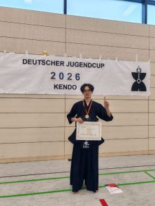 Ryusei Aoki vor dem Banner des Deutschen Jugendcup 2026 mit Urkunde für den ersten Platz